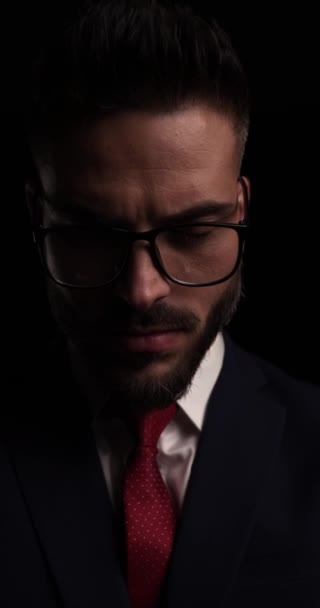 vídeo vertical de belo jovem empresário vestindo terno e óculos olhando para o lado e posando em fundo preto - Filmagem, Vídeo