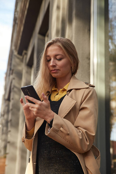 donna caucasica in trench beige con smartphone all'aperto nella giornata di sole - Foto, immagini
