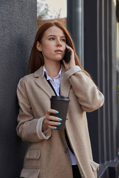 белая женщина в стильном бежевом пальто звонит по смартфону с чашкой кофе - Фото, изображение