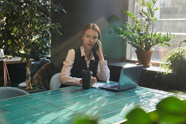 студентка, яка сидить за столом кафе, навчається з ноутбуком, телефонує на смартфон
 - Фото, зображення