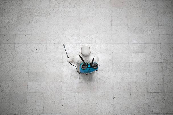 Widok osoby nierozpoznawalnej w białym kombinezonie ochrony chemicznej, dezynfekującej miejsca publiczne, aby powstrzymać rozprzestrzenianie się wysoce zaraźliwego wirusa Covid-19. - Zdjęcie, obraz