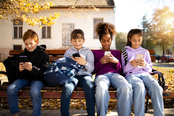 Groupe d'enfants multiculturels assis sur un banc et utilisant leurs téléphones intelligents surfer sur Internet concept de technologie. - Photo, image