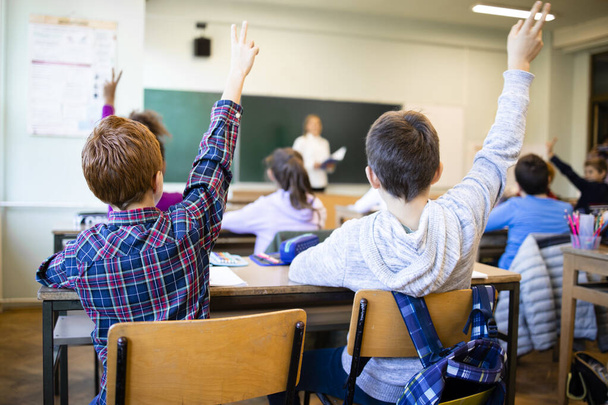 Escolares em sala de aula com as mãos levantadas respondendo à pergunta do professor. - Foto, Imagem