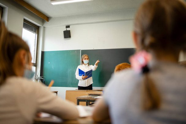 Lehrerin während Coronavirus-Pandemie trägt Mundschutz und unterrichtet Schüler im Klassenzimmer. - Foto, Bild