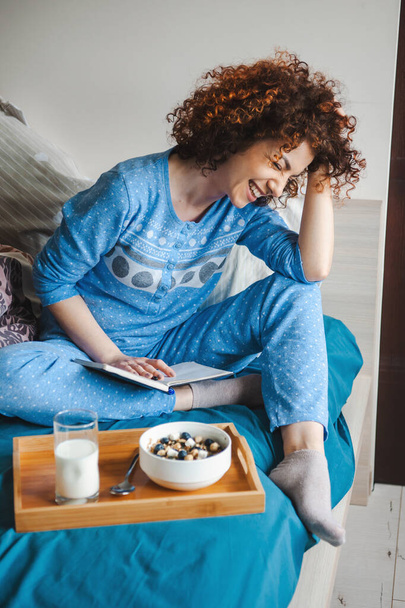 Портрет усміхненої молодої жінки, що сидить у блакитній піжамі на ліжку, читає цікаву книгу. Сніданок у ліжку. Час у ліжку. кучеряве волосся
. - Фото, зображення