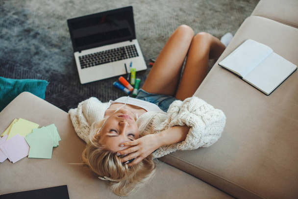 Bovenaanzicht van een blanke vrouw zittend op de vloer met haar hoofd leunend op de bank moe van zoveel studie. Online onderwijs. - Foto, afbeelding