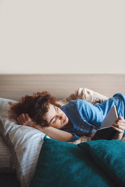 Kręcona kobieta leżąca na łóżku w niebieskiej piżamie i czytająca książkę. Dzień dobry. Czas do łóżka. Koncepcja stylu życia. - Zdjęcie, obraz
