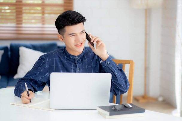Jonge Aziatisch man praten telefoon en werken vanuit huis met laptop computer, freelance man schrijven op notebook en spreken op smartphone, thuis blijven, huishoudelijk leven, zakelijke en communicatie concept. - Foto, afbeelding