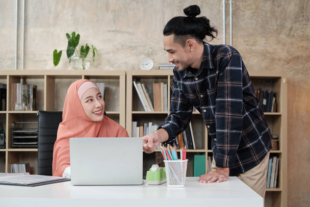 Dva mladí začínající kolegové, kteří jsou islámskými lidmi, hovoří s úsměvem o úspěchu v elektronickém obchodu. Použití notebooku pro on-line komunikaci přes internet v malé kanceláři. - Fotografie, Obrázek
