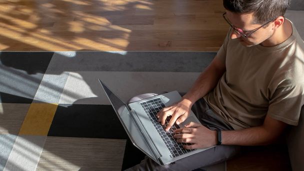 Baner młodego mężczyzny w okularach siedzącego na podłodze w nowoczesnym i stylowym wnętrzu i pracującego z laptopem - Zdjęcie, obraz