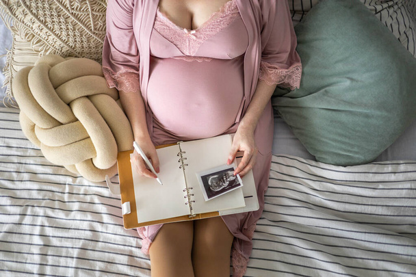 Top view caring future mother writing diary with ultrasound x ray photo sitting on comfortable bed - 写真・画像