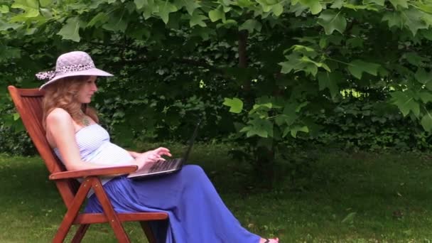 Беременная женщина с ноутбуком в шляпе сидит на стуле в зеленом парке
 - Кадры, видео