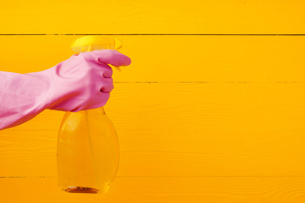 Hand in glove holding household detergent spray against yellow wooden background - Foto, immagini