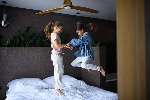 Two little sister holding hands and jumping on bed indoors in hotel. - Foto, Imagen
