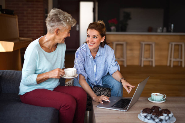 Senior mother having coffee with adult daughter indoors at home, sitting, talking and using tablet. - Photo, Image