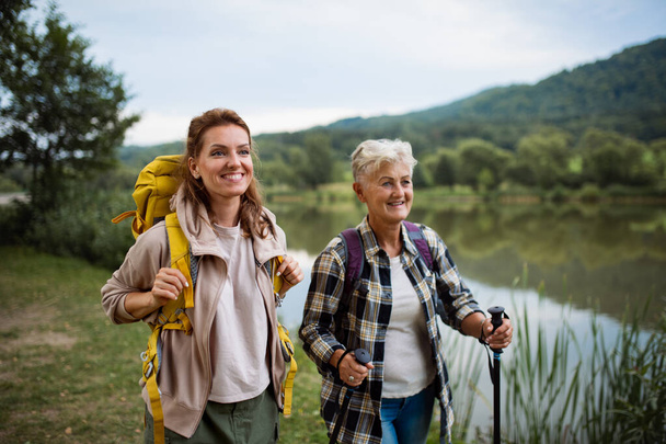 Happy senior woman with trekking poles hiking with adult daughter outdoors in nature. - Fotó, kép