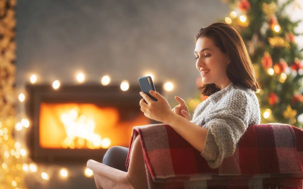 Mujer está utilizando el teléfono móvil sentado cerca del árbol de Navidad. - Foto, imagen