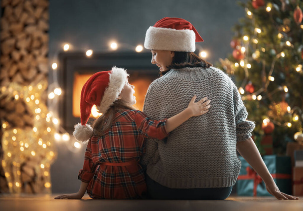 Merry Christmas and Happy Holidays. Mom and her cute daughter girl looking at fireplace. Parent and little child having fun near tree indoors. - Фото, изображение