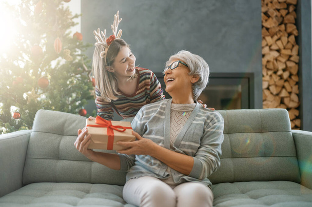 Merry Christmas and Happy Holidays. Cheerful grandma and her cute grand daughter exchanging gifts. Granny and child having fun near tree indoors. Loving family with presents in room. - Zdjęcie, obraz
