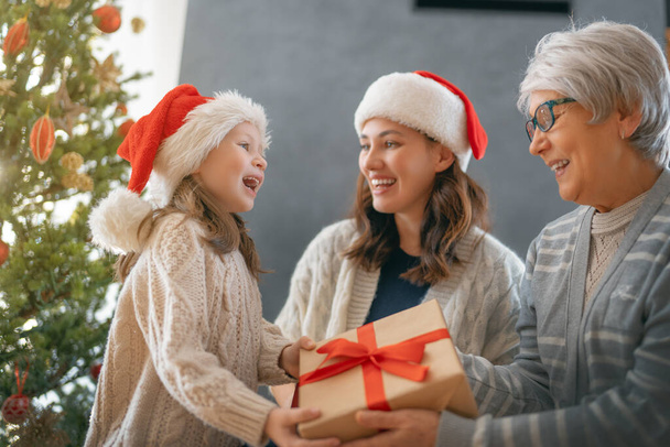 Merry Christmas and Happy Holidays. Cheerful kid presenting gifts to mom and granny. Parents and little child having fun near tree indoors. Loving family with presents in room. - 写真・画像