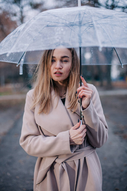 A young, slender, beautiful girl in a coat walks in the rain with a transparent umbrella in the park - Фото, зображення