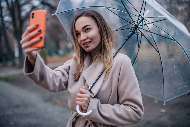 Chica joven feliz con un paraguas transparente toma una selfie en un día lluvioso - Foto, Imagen