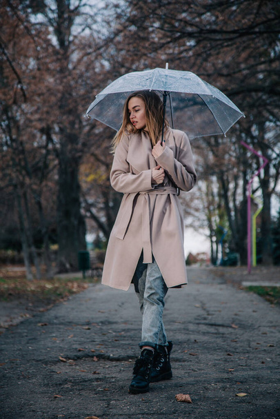 A young, slender, beautiful girl in a coat walks in the rain with a transparent umbrella in the park - Photo, image