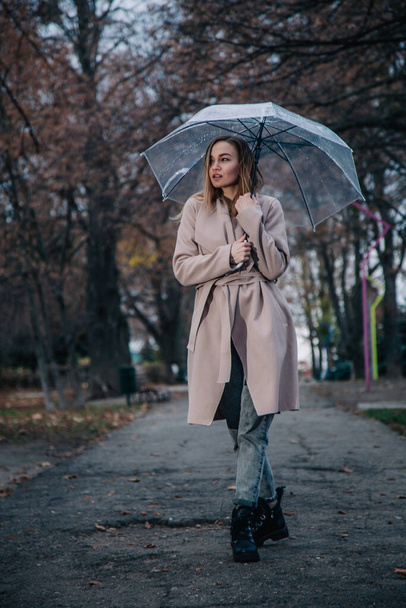 Mladá, štíhlá, krásná dívka v kabátě kráčí v dešti s průhledným deštníkem v parku - Fotografie, Obrázek
