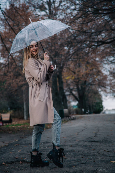A young, slender, beautiful girl in a coat walks in the rain with a transparent umbrella in the park - Фото, изображение