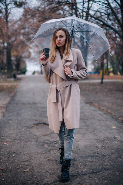 A young, beautiful girl with a disposable cup of coffee walks in the park under a transparent umbrella in the rain - Фото, изображение