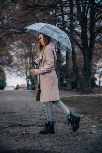 A young, slender, beautiful girl in a coat walks in the rain with a transparent umbrella in the park - Fotoğraf, Görsel