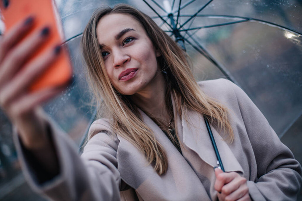 Happy young girl with a transparent umbrella takes a selfie on a rainy day - Фото, зображення
