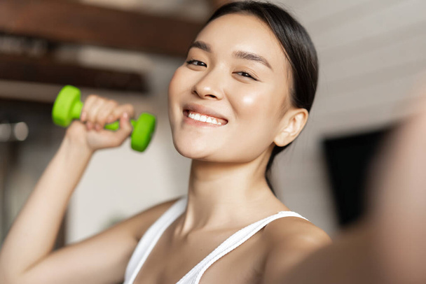 Portrait d'instructeur de fitness asiatique souriant prenant selfie avec haltère. Femme athlétique faisant photo sur téléphone portable avec équipement de sport, entraînement à la maison - Photo, image