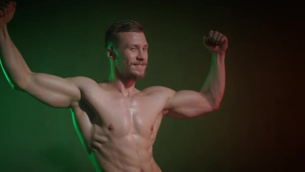 Egy csupasz testű sportoló áll egy pózban, hogy megmutassa a bicepszét. Mosolyog és a kamerába néz. Narancssárga és zöld fény világít rá. 4K - Felvétel, videó