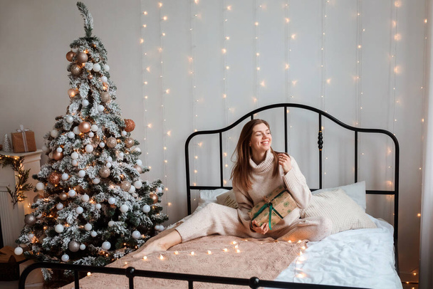 Romantic woman is sitting on the bed with Christmas present in her hands, next to Christmas tree. Girl spending winter holidays at home. - Fotoğraf, Görsel
