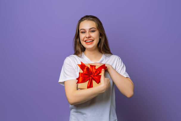 Foto van gelukkige jonge brunette vrouw in casual wit t-shirt met geschenkdoos en kijkend naar de camera, geïsoleerd staande over paarse achtergrond. Nieuwjaar Vrouwendag verjaardagsvakantie concept - Foto, afbeelding
