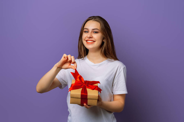 Gelukkig glimlachende jonge brunette vrouw in casual wit t-shirt opening verpakt cadeau. Nieuwjaar Vrouwendag verjaardagsvakantie concept - Foto, afbeelding