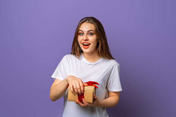 Omg. Pretty girl wearing casual t shirt posing at purple blank wall with opened box, keeping jaw dropped, looking in full disbelief, receiving best ever present on New Year - Foto, afbeelding