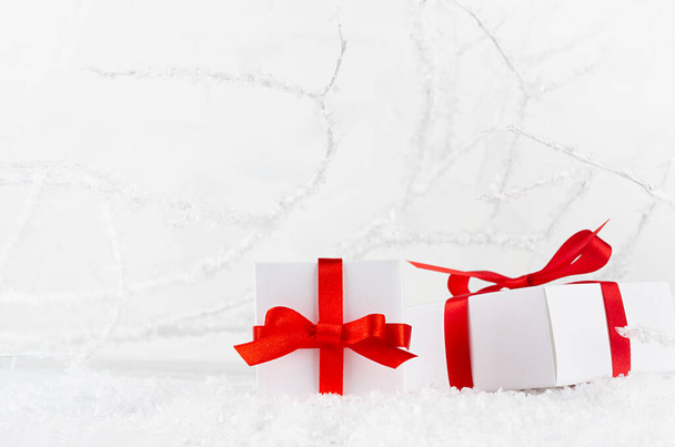 Dos cajas de regalo festivas de color blanco brillante con primer plano de cinta de satén rojo en el bosque de invierno decorativo con ramas heladas y nieve, espacio para copiar. Fondo navideño con embalaje navideño para celebración. - Foto, imagen