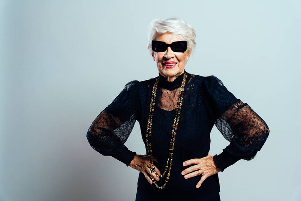 Beautiful and elegant old influencer woman. Cool grandmother posing in studio wearing fashionable clothes. Happy senior lady celebrating and making party. Concept about seniority and lifestyle - Foto, Imagem
