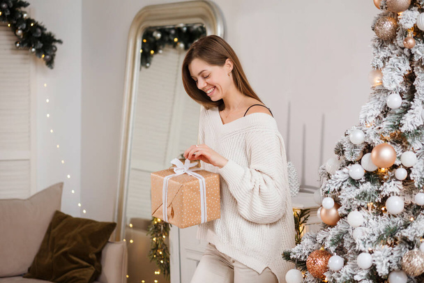 Smiling woman is opening present box standing next to Christmas tree, indoors.  - Foto, immagini