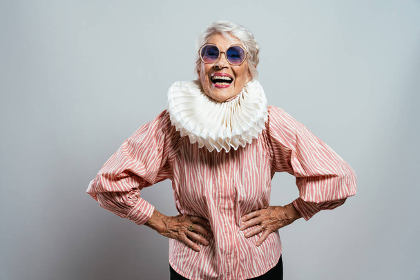 όμορφη και κομψή γριά γυναίκα με επιρροή. Ωραία γιαγιά να ποζάρει στο στούντιο φορώντας μοντέρνα ρούχα. Ευτυχισμένη ηλικιωμένη κυρία γιορτάζει και κάνει πάρτι. Έννοια για την αρχαιότητα και τον τρόπο ζωής - Φωτογραφία, εικόνα