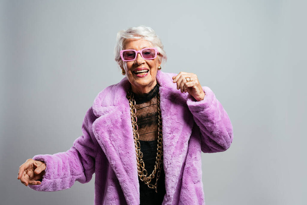 image of a beautiful and elegant old influencer woman. Cool grandmother posing in studio wearing fashionable clothes. Happy senior lady celebrating and making party. Concept about seniority and lifestyle - Photo, image