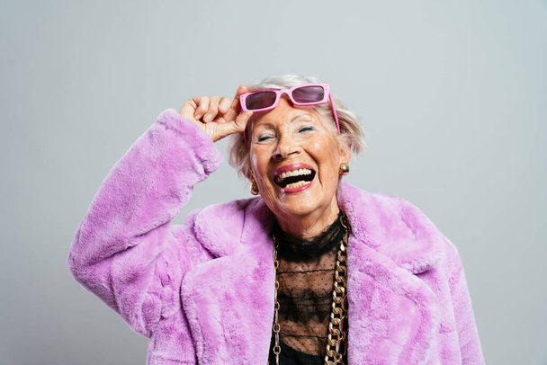 image of a beautiful and elegant old influencer woman. Cool grandmother posing in studio wearing fashionable clothes. Happy senior lady celebrating and making party. Concept about seniority and lifestyle - Foto, Imagem