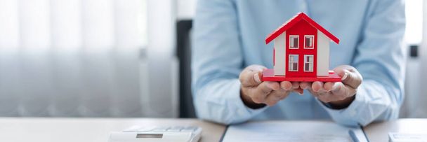 Das Immobilienkonzept, mit dem der Makler seinem Kunden seine Arbeit präsentiert, indem er das rote Hausmodell auf die Handflächen legt und ein wenig die Hände hebt. - Foto, Bild