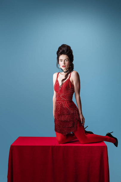 Portrait of young adorable woman in dark red, wine color evening dress posing on table with red cloth isolated on blue background. - Foto, afbeelding