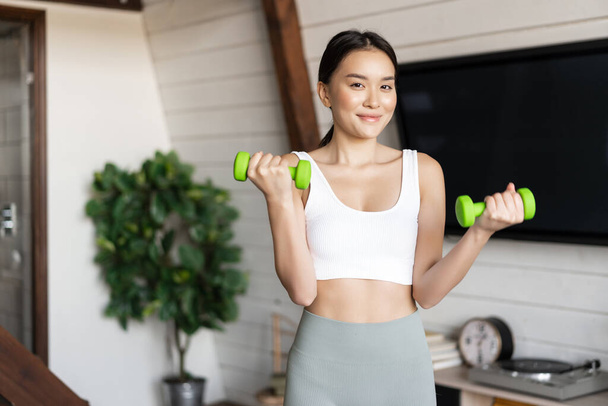 Fitness aziatische meisje doet workout thuis in de woonkamer, tillen halters, het dragen van activewear voor sport activiteiten - Foto, afbeelding