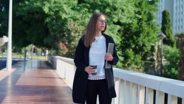 Imprenditrice donna in elegante abbigliamento con computer portatile a piedi in città e utilizzando il cellulare sul ponte - Filmati, video