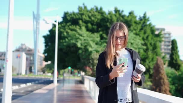 Grave imprenditrice in abito elegante con laptop che cammina in città e parla al cellulare mentre beve un caffè - Filmati, video