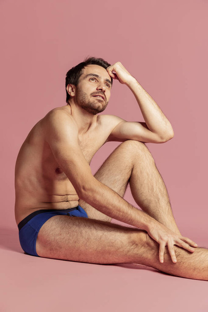 Studio shot of young handsome bearded shirtless man in blue panties sitting on floor isolated on pink background. - Fotografie, Obrázek